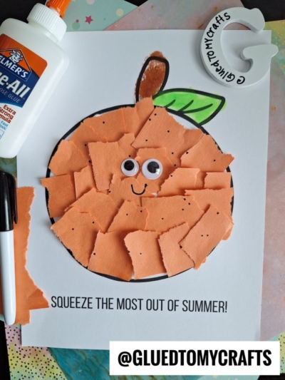 A torn paper orange craft features googly eyes and a drawn smile on white paper. Nearby are a glue bottle, marker, and a GLUEDTOMYCRAFTS sticker, making this playful creation stand out.