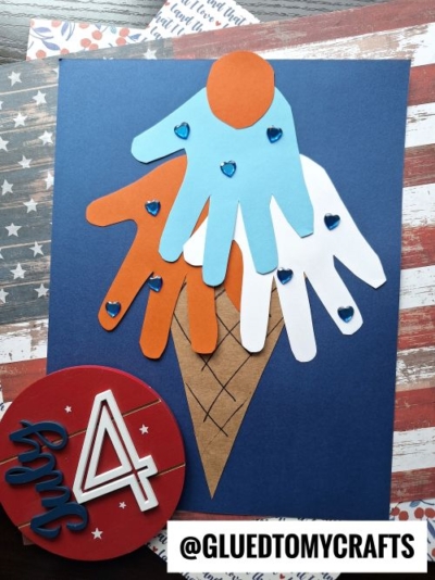 A craft depicting an ice cream cone made from handprint cutouts in blue, orange, and white paper on a navy background, decorated with blue heart stickers—perfect for 4th of July keepsakes, with patriotic paper featured in the background.