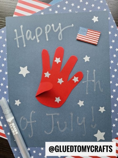 A blue paper with Happy 4th of July! written in silver, decorated with white stars, a red handprint cutout, a small American flag sticker, and a marker, atop red and white striped paper—perfect for creating memorable 4th of July keepsakes.