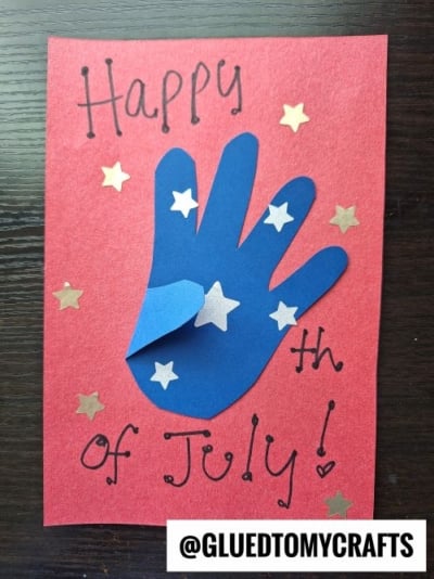 A red card with Happy 4th of July! written on it, decorated with a blue handprint cutout and white stars—perfect for 4th of July keepsakes. The card is shown on a dark surface with the handle @GLUEDTOMYCRAFTS at the bottom.