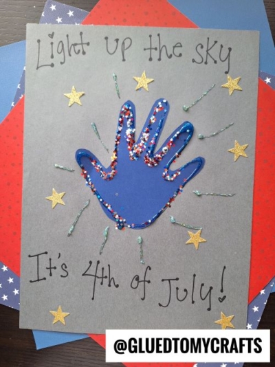 A handmade 4th of July card features a blue handprint outlined with glitter, surrounded by gold stars on gray paper. Perfect for 4th of July keepsakes, it reads Light up the sky It’s 4th of July! and rests atop colorful papers.
