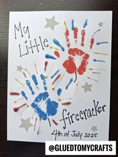 A white paper features red and blue handprints with gold streaks, silver stars, and the text My Little firecracker 4th of July 2025 in playful fonts—perfect for 4th of July keepsakes. The handle @GLUEDTOMYCRAFTS appears at the bottom.