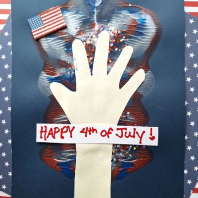 A handprint cutout is glued to dark paper with red, white, and blue paint, star-shaped glitter, a mini American flag, and a “Happy 4th of July!” sign—perfect for making memorable 4th of July keepsakes. The background features patriotic striped and starred paper.