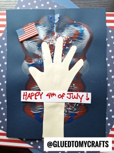 A handprint cutout is glued to dark paper with red, white, and blue paint, star-shaped glitter, a mini American flag, and a “Happy 4th of July!” sign—perfect for making memorable 4th of July keepsakes. The background features patriotic striped and starred paper.