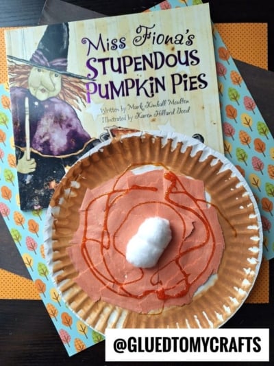 A children’s book titled “Miss Fiona’s Stupendous Pumpkin Pies” sits beside a torn paper pie craft, decorated with orange paint, brown paper, and a cotton ball “whipped cream.” The bottom reads @GLUEDTOMYCRAFTS.