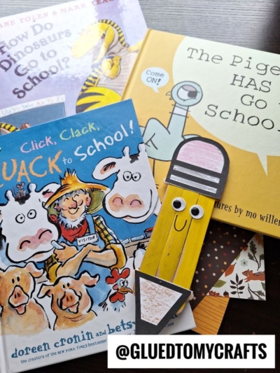 A collection of children’s books about school, with a cute popsicle stick pencil bookmark with googly eyes in the foreground. A @GLUEDTOMYCRAFTS label appears at the bottom of the image.