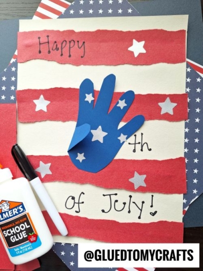 A handmade Fourth of July card with red stripes, white stars, and a blue paper handprint makes the perfect 4th of July keepsake. Supplies like glue and a marker are nearby. The card says Happy 4th of July! with @gluedtomycrafts at the bottom.