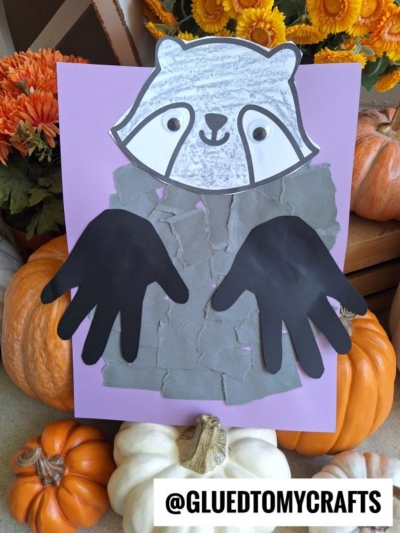 A torn paper raccoon craft featuring a raccoon face, a gray paper body, and black handprint cutout arms, displayed among pumpkins and flowers.
