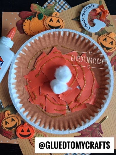 A torn paper pie craft using a paper plate to resemble pumpkin pie, featuring cotton as whipped cream, orange torn paper filling, glitter accents, and pumpkin-themed decorations on a fall background. Glue and a craft tag are also shown.
