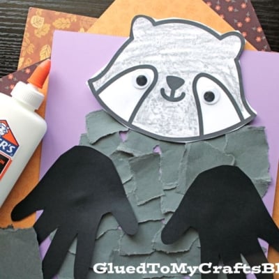 A torn paper raccoon craft made with grey paper for the body, black handprint arms, and a cute raccoon face, all displayed on purple paper beside Elmer’s glue.