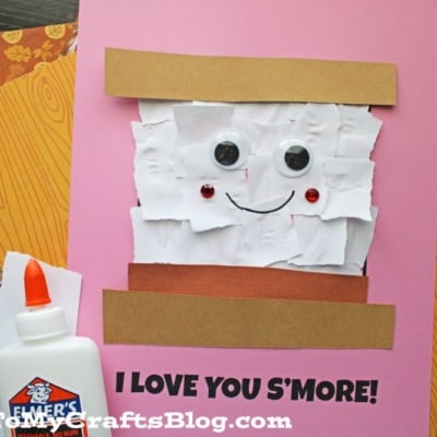 A pink card with torn paper smores details—white paper, googly eyes, and a smile—resembles a s'more with “I LOVE YOU SMORE!” written below. Brown strips act as graham crackers.