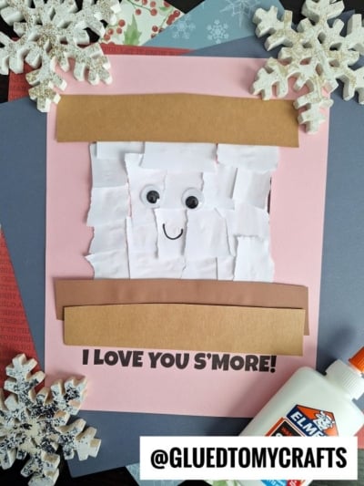 A pink card featuring a torn paper s’more with googly eyes and a smile. "I LOVE YOU S’MORE!" is printed at the bottom. Snowflake decorations, glue, and craft paper surround the cute torn paper smores creation.