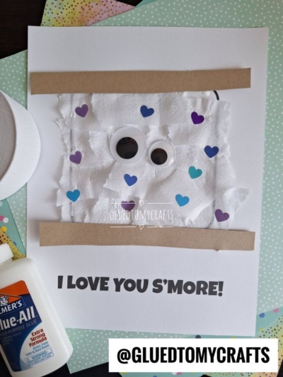 A craft project on white paper shows a googly-eyed, torn paper smores with tissue and heart stickers, brown strips for graham crackers, and the text I LOVE YOU SMORE! Glue and craft supplies are nearby.