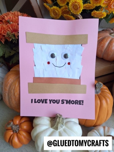 A pink card featuring a torn paper smores design with googly eyes sits among pumpkins and flowers. The card says, I LOVE YOU SMORE! and has the handle @GLUEDTOMYCRAFTS at the bottom.