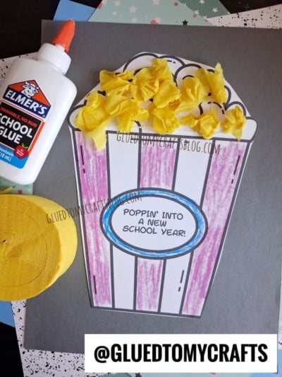 A fun back to school popcorn craft featuring yellow tissue popcorn, a glue bottle, and a roll of yellow streamers. The paper popcorn container reads "Poppin into a new school year!" on a gray background—perfect for celebrating the start of the school year with some creative flair.
