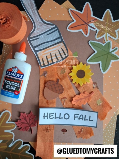 Craft project with fall-themed paper cutouts, orange tissue paper, Elmers glue, and a painted paper brush as part of a charming fall paint brush craft. A HELLO FALL sign is arranged on a polka dot background, with @GLUEDTOMYCRAFTS visible at the bottom.