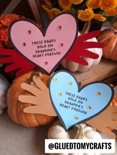 Two handmade heart crafts with cut-out handprints and sweet “hold my heart” messages for Grandma and Grandpa, perfect for Grandparent's Day, surrounded by pumpkins and flowers.