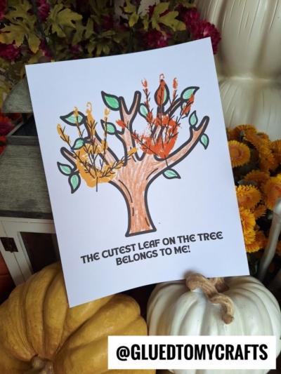 A handprint autumn tree craft with the text The cutest leaf on the tree belongs to me! sits among pumpkins, flowers, and fall décor—making it a perfect cutest leaf keepsake for the season. The username @GLUEDTOMYCRAFTS appears at the bottom right.