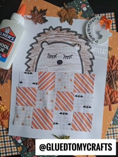 A sleeping hedgehog craft is displayed on a table, featuring squares of patterned paper, a bottle of Elmer’s glue, faux autumn leaves, and orange and brown paper. The text @GLUETOMYCRAFTS is visible on the image.