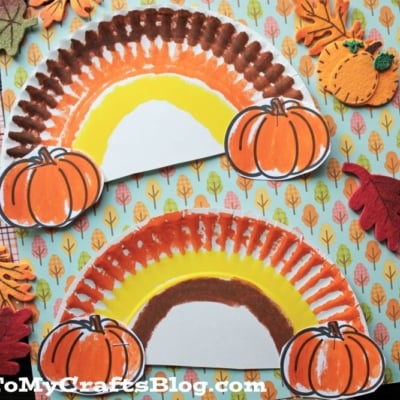 Two paper plate crafts painted as Paper Plate Fall Rainbow in autumn hues, each adorned with pumpkin cutouts, are displayed on seasonal leaf-themed paper.