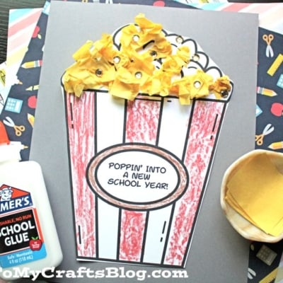 A back to school popcorn craft features a popcorn bucket with yellow tissue paper popcorn pieces, decorated and labeled Poppin Into a New School Year! Glue, a cup, and printed paper are nearby.