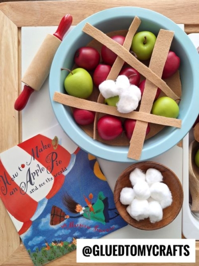 A fall themed sensory bin idea: a play food setup with red and green toy apples in a blue pie dish, topped with brown felt strips and cotton balls, next to a children’s book and a bowl of cotton balls.