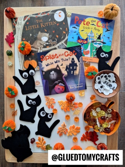 A fall-themed craft setup with black cat cutouts, googly eyes, fake leaves, small pumpkins, autumn flowers, and three Halloween cat books on a tray—perfect for inspiring FALL THEMED SENSORY BIN IDEAS. @GLUEDTOMYCRAFTS is written at the bottom.
