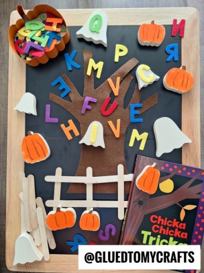 A Halloween-themed craft setup with a felt tree, foam letters, pumpkins, ghosts, a craft stick fence, and the book Chicka Chicka Boom Boom—perfect inspiration for FALL THEMED SENSORY BIN IDEAS. Supplies are neatly arranged on a black tray.