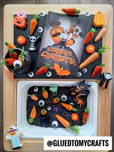 A Halloween-themed sensory tray with fake carrots, toy eyeballs, spiders, and Halloween character cutouts inspired by the Creepy Carrots! bookāperfect for anyone looking for fun fall themed sensory bin ideas, all arranged on a wooden surface.