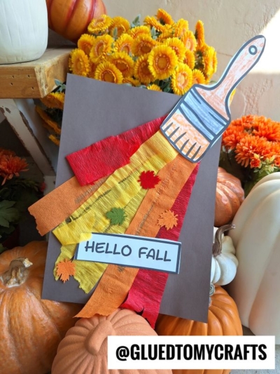 A fall paint brush craft features a paper paintbrush “painting” autumn colors with crepe paper strips and leaf cutouts on brown paper, accented by a “HELLO FALL” sign, pumpkins, and orange flowers.