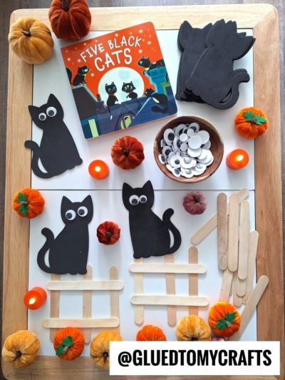 A Halloween craft setup with black cat cutouts, googly eyes, popsicle sticks, felt pumpkins, LED candles, and the book Five Black Cats on a wooden tray—perfect inspiration for FALL THEMED SENSORY BIN IDEAS. @GLUEDTOMYCRAFTS is written at the bottom.