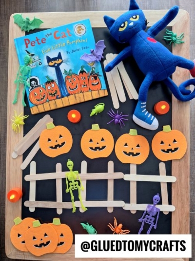 A flat lay of Halloween crafts inspired by Pete the Cat: Five Little Pumpkins, featuring a blue plush cat, popsicle stick fences, paper pumpkins, toy skeletons, spiders, and LED tea lights—perfect for fall themed sensory bin ideas.