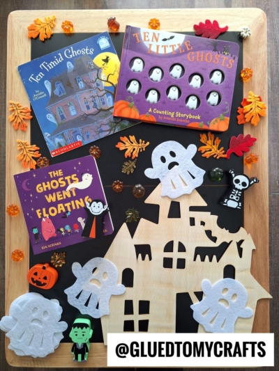 A wooden tray displays Halloween-themed children’s books, felt ghost and pumpkin decorations, small toy figures, artificial autumn leaves, and a haunted house cutout—perfect inspiration for FALL THEMED SENSORY BIN IDEAS. The @gluedtomycrafts handle appears at the bottom.
