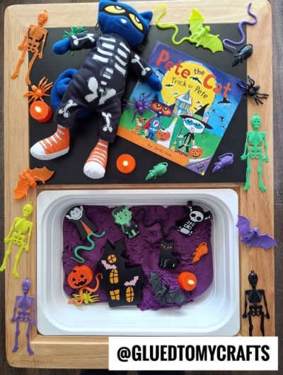 A fall themed sensory bin idea, this kids’ Halloween activity tray features purple kinetic sand, plastic skeletons, bats, spiders, a black cat, pumpkin cutouts, plush Pete the Cat in a skeleton costume, and the book “Pete the Cat: Trick or Pete.”.