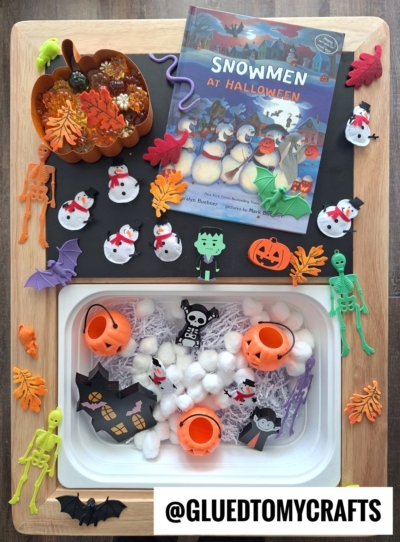 A Halloween-themed sensory play tray inspired by FALL THEMED SENSORY BIN IDEAS, featuring pumpkin and skeleton decorations, foam snowmen, bats, fake spiders, cotton balls, plastic pumpkins, and the book Snowmen at Halloween. @GLUEDTOMYCRAFTS is visible.
