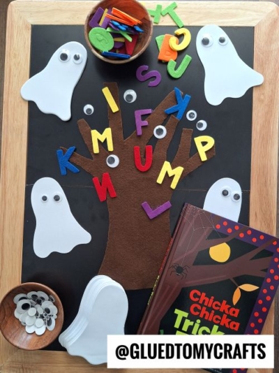 A Halloween-themed craft setup with a felt tree, foam letters, googly eyes, paper ghosts, and the book Chicka Chicka Trick or Treat on a tray—perfect for inspiring FALL THEMED SENSORY BIN IDEAS. The @GLUEDTOMYCRAFTS watermark is visible.