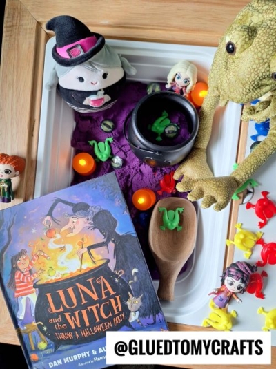 A perfect pick for FALL THEMED SENSORY BIN IDEAS, this Halloween bin features purple play dough, a toy cauldron, lights, a plastic witch, dinosaur hand, small figures, and the book Luna and the Witch. Tag: @GLUEDTOMYCRAFTS.