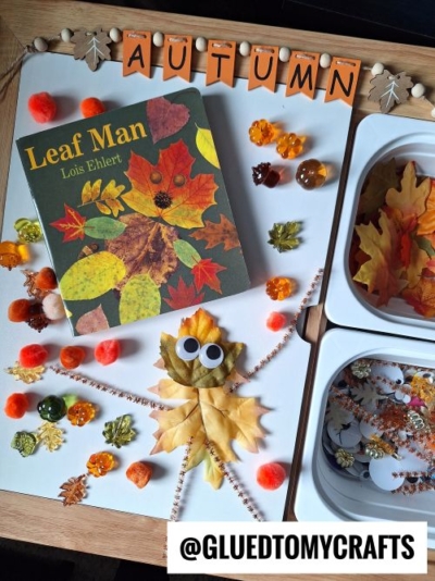 A fall-themed craft setup with the book Leaf Man by Lois Ehlert, fake leaves, pom-poms, gems, and a leaf man made from leaves and googly eyes—perfect inspiration for your FALL THEMED SENSORY BIN IDEAS. "AUTUMN" is spelled out in flags above the display.