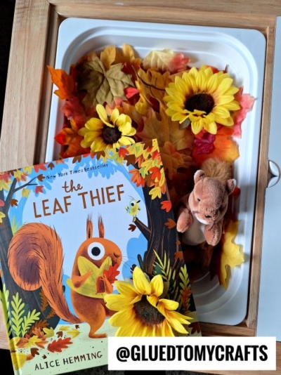 A children's book titled The Leaf Thief is placed on a tray decorated with artificial sunflowers, autumn leaves, and a small plush squirrel—perfect inspiration for FALL THEMED SENSORY BIN IDEAS. The Instagram handle @GLUEDTOMYCRAFTS is visible at the bottom.