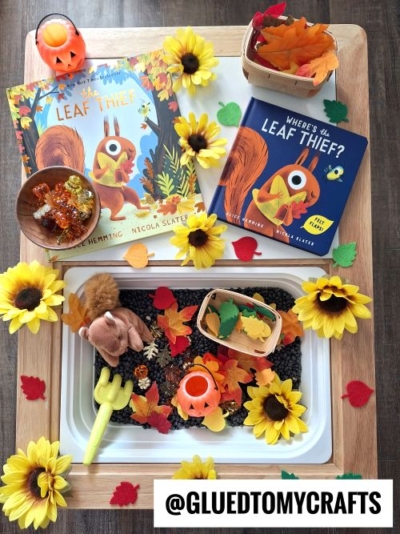 A cozy fall-themed sensory bin setup featuring sunflowers, colorful fake leaves, black beans, small pumpkins, a plush squirrel, honey sticks, and two versions of Where’s My Leaf Thief?—perfect for inspiring new FALL THEMED SENSORY BIN IDEAS.