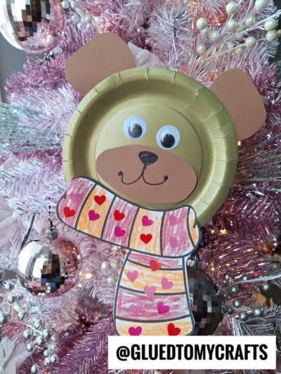 A paper plate bear face made from construction paper, with googly eyes and a colorful scarf decorated with hearts, hangs on a pink and white Christmas tree. Text reads @GLUEDTOMYCRAFTS.