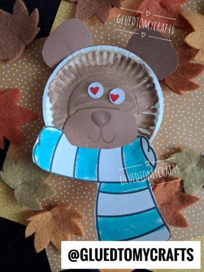 A cute lion face made from a painted paper plate, with paper ears, nose, and scarf—similar in style to a paper plate bear—features heart-shaped eyes on a dotted background with paper leaves. Text: @GLUEDTOMYCRAFTS.