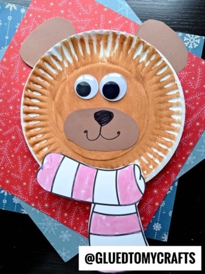 A paper plate bear face with paper ears, googly eyes, a paper nose and mouth, plus a drawn pink-and-white striped scarf on red and blue snowflake backgrounds. A label reads “@GLUEDTOMYCRAFTS.”.
