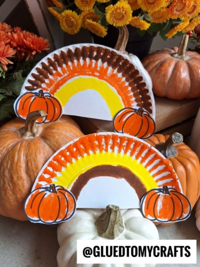 Two Paper Plate Fall Rainbow crafts, painted in autumn shades and adorned with paper pumpkins, are displayed among real pumpkins and orange flowers. The text @GLUEDTOMYCRAFTS appears at the bottom of the image.