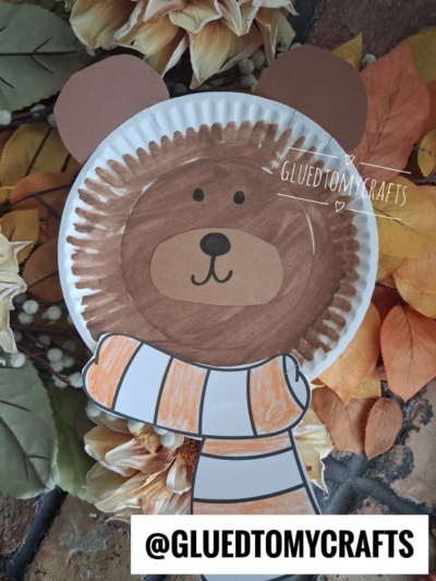 A charming paper plate bear craft with brown fur, round ears, and a white-and-orange scarf lies on autumn leaves and flowers. @GLUEDTOMYCRAFTS is written at the bottom.