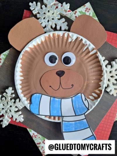 A paper plate bear face made from brown and white plates, with brown paper ears, googly eyes, a paper nose and mouth, plus a blue-striped scarf. Snowflake decorations surround this adorable craft. @GLUEDTOMYCRAFTS is labeled at the bottom.