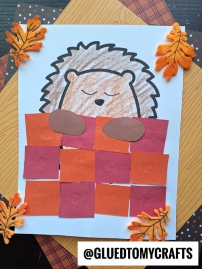 A drawing of a sleeping hedgehog craft tucked under a blanket made of red and orange paper squares, with felt leaves decorating the corners. The image has a gluedtomycrafts watermark at the bottom.