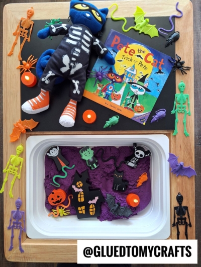 A Halloween sensory tray inspired by FALL THEMED SENSORY BIN IDEAS includes a Pete the Cat book, a skeleton-costumed doll, spooky toys like bats and spiders, purple sand, and plastic skeletons. The @GLUEDTOMYCRAFTS handle is also visible.