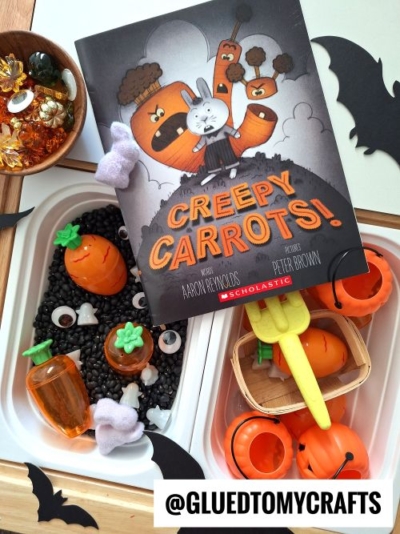 Childrenās book āCreepy Carrots!ā is displayed on a sensory play tray filled with black beans, toy carrots, pumpkins, scoops, and Halloween decorāperfect inspiration for fall themed sensory bin ideas. @GLUEDTOMYCRAFTS watermark visible.