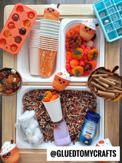 A tabletop with fall-themed craft supplies—orange cups, pumpkin decorations, felt pumpkins, cinnamon sticks, ice cube trays, coffee creamer, and shredded paper—perfect for inspiring FALL THEMED SENSORY BIN IDEAS in beautiful autumn colors. Tag: @GLUEDTOMYCRAFTS.